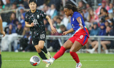 Jaedyn Shaw kicks a ball while wearing a blue and red national team kit. Ji So-yun runs behind her.